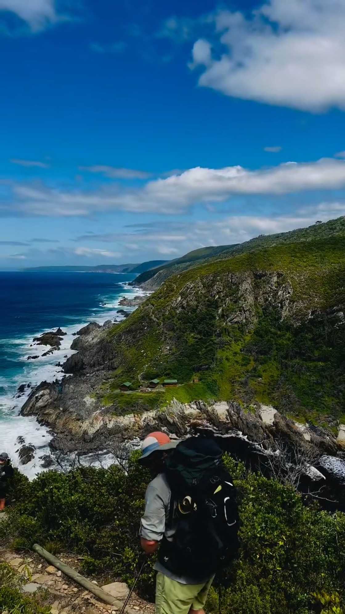 A hiker with a large backpack walks along a narrow trail above a rugged coastline, with steep green cliffs and waves crashing against the rocks under a blue sky.