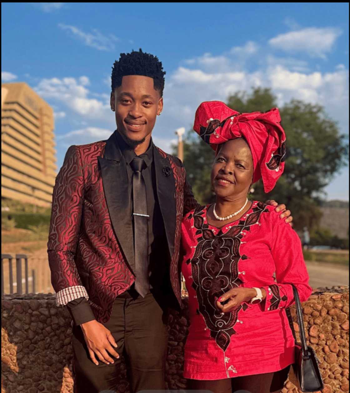 Graduate standing next to a woman dressed in vibrant traditional attire, both posing outdoors against a backdrop of buildings and trees.