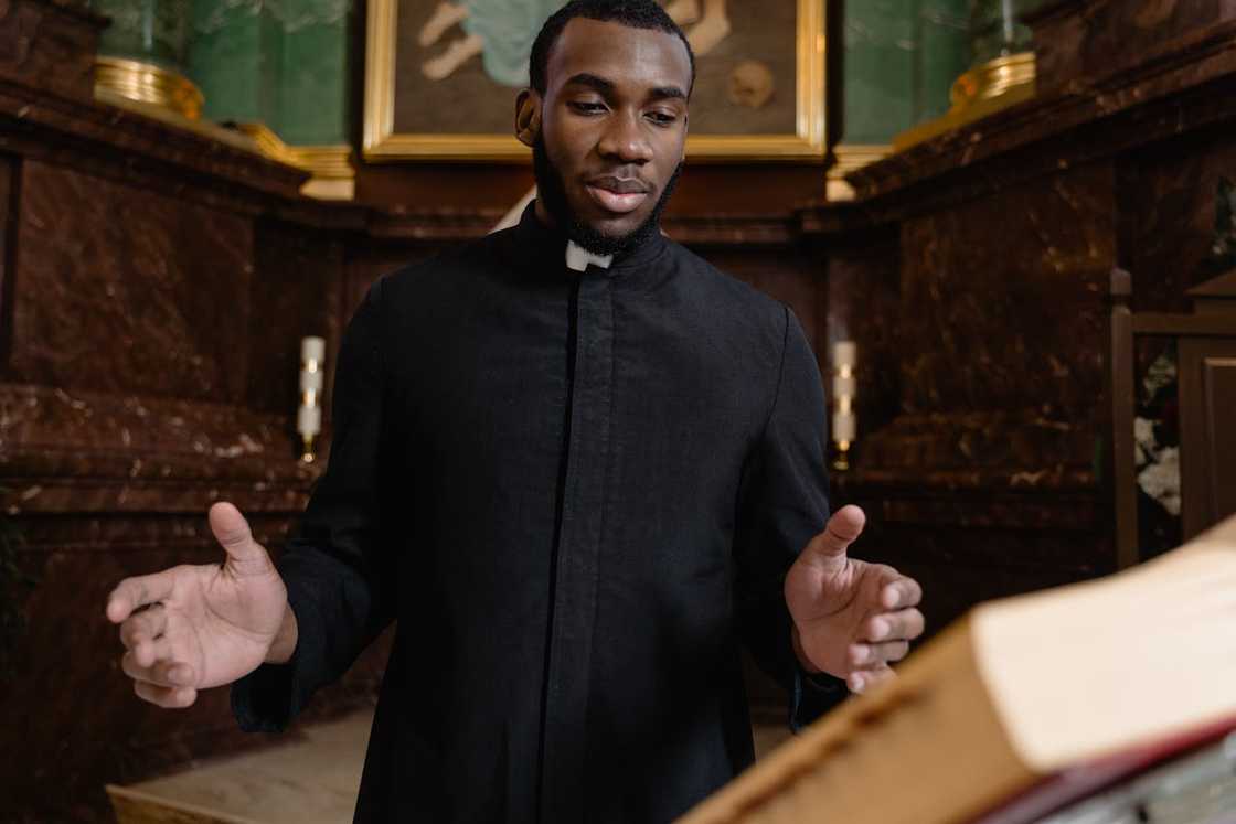 A man in clerical robe standing indoors with hands raised in a prayerful gesture. A man in clerical robe standing indoors with hands raised in a prayerful gesture.