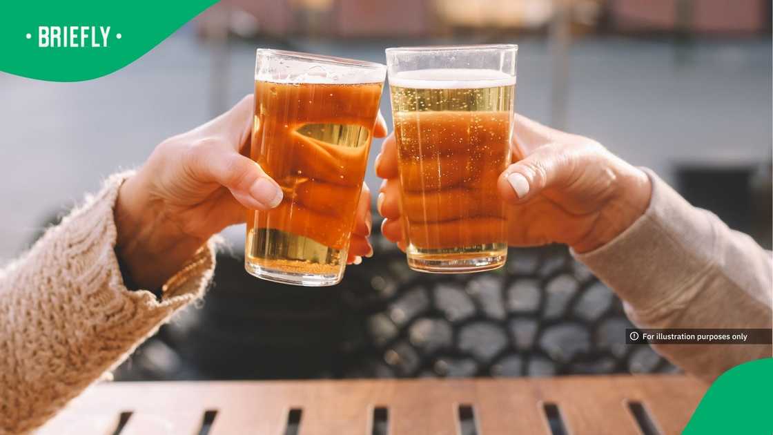 Close-up of two people toasting with pint glasses of beer while sitting at an outdoor wooden table