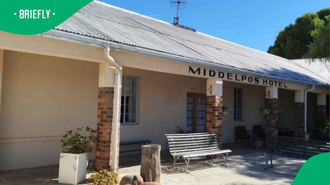 The front of the Middelpos Hotel appeared quiet and sunlit, with simple outdoor seating and a traditional structure surrounded by greenery