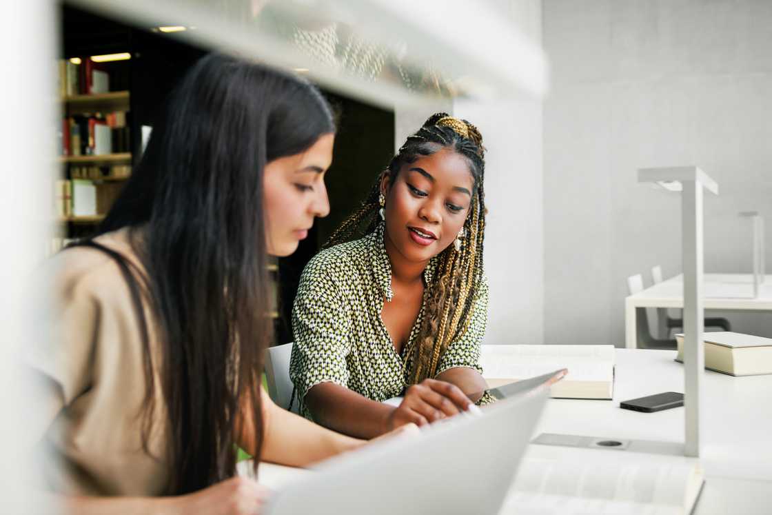 Two students are engaged in a discussion while referring to a laptop and textbooks. Two students are engaged in a discussion while referring to a laptop and textbooks.