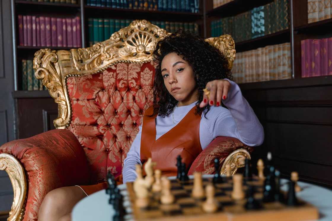 A woman in a red and gold armchair, holding a chess piece in a library setting. A woman in a red and gold armchair, holding a chess piece in a library setting.