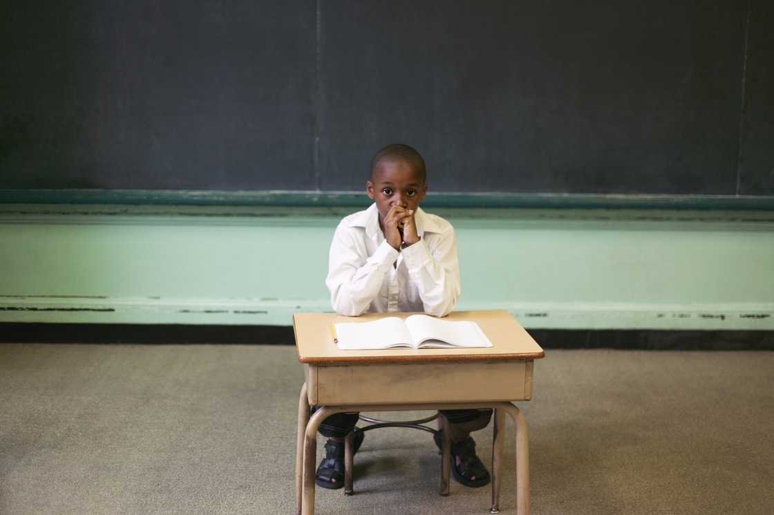 Child sits alone at a small desk in front of a blank notebook, hands clasped near face. Child sits alone at a small desk in front of a blank notebook, hands clasped near face.