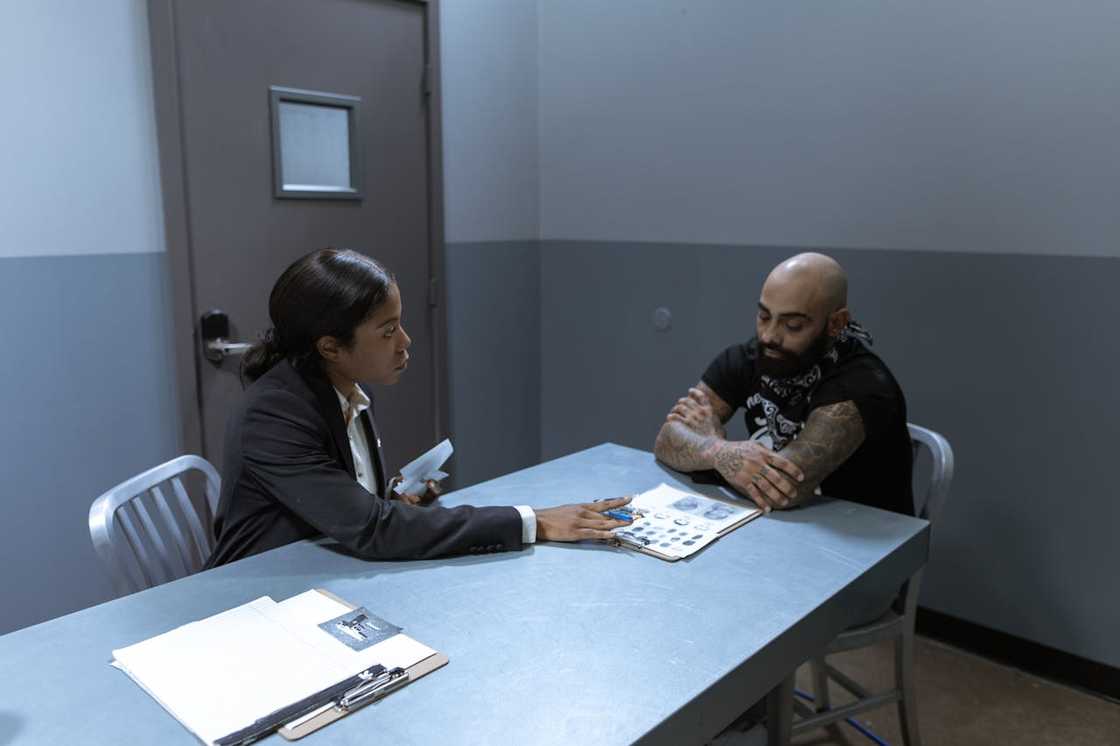 A detective interviews a tattooed suspect at a metal table.