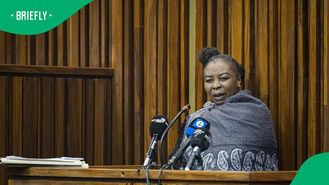 Convicted former police officer Nomia Rosemary Ndlovu stands inside the prisoner's dock in a South African courtroom, wearing a colorful patterned top, styled hair, and a dark face mask lowered beneath her chin.