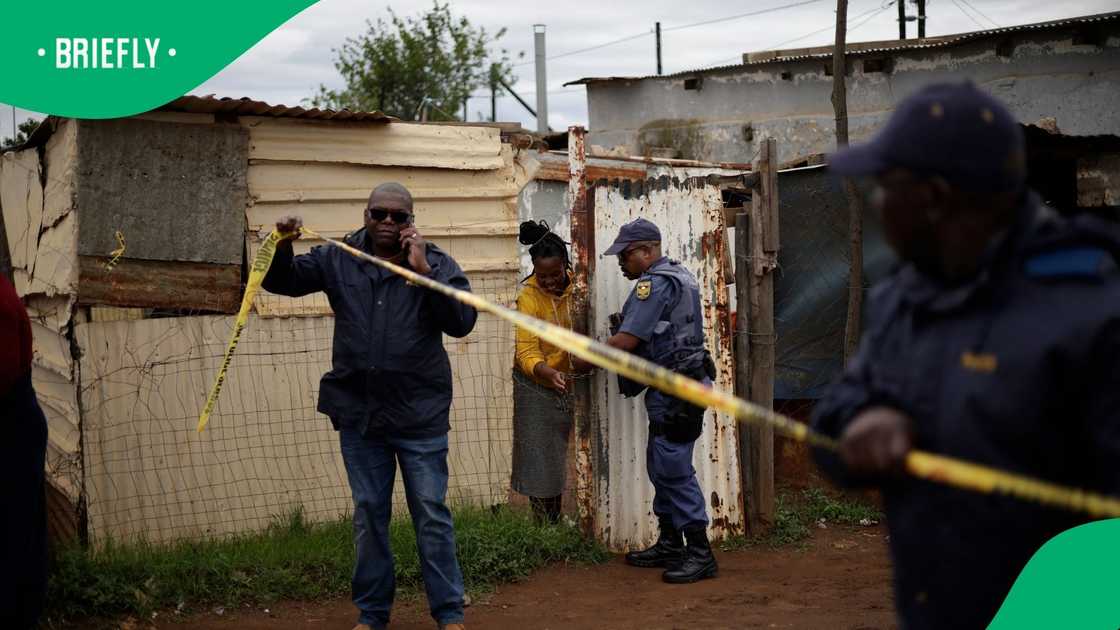 South African Police Service officers standing near yellow crime scene tape that blocks off a dirt road in a township, while a detective in civilian clothes speaks with a resident outside a brick home.