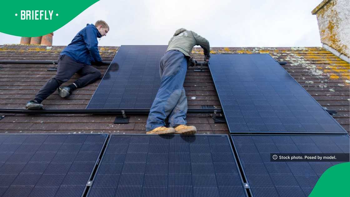 One individual leaned over a large, dark solar panel to secure it while their teammate maintained their balance on the upper portion of the roof.