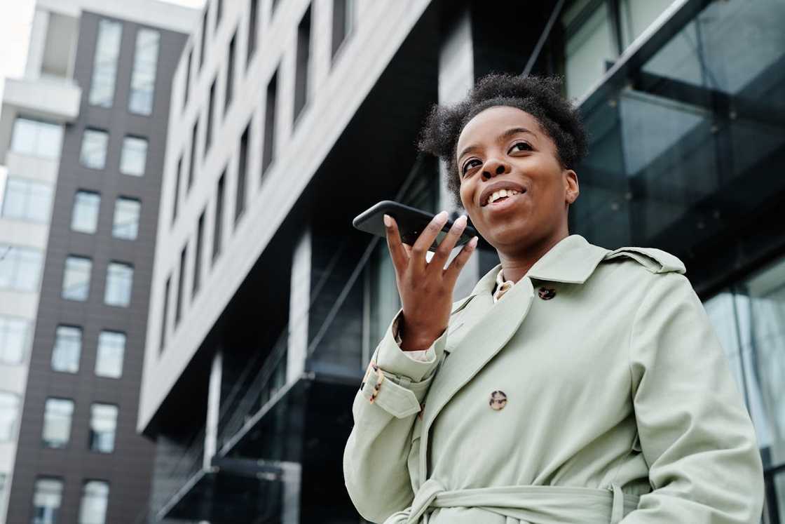 A woman speaks into a smartphone outside a modern office building.