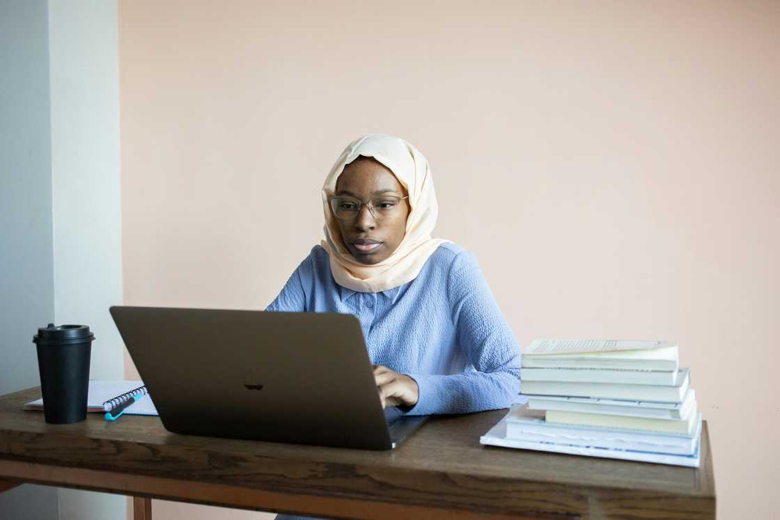 A serious young lady in a hijab and a blue top is using a laptop