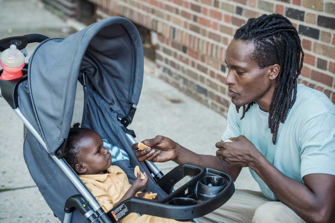A father feeds a baby seated in a stroller outdoors.