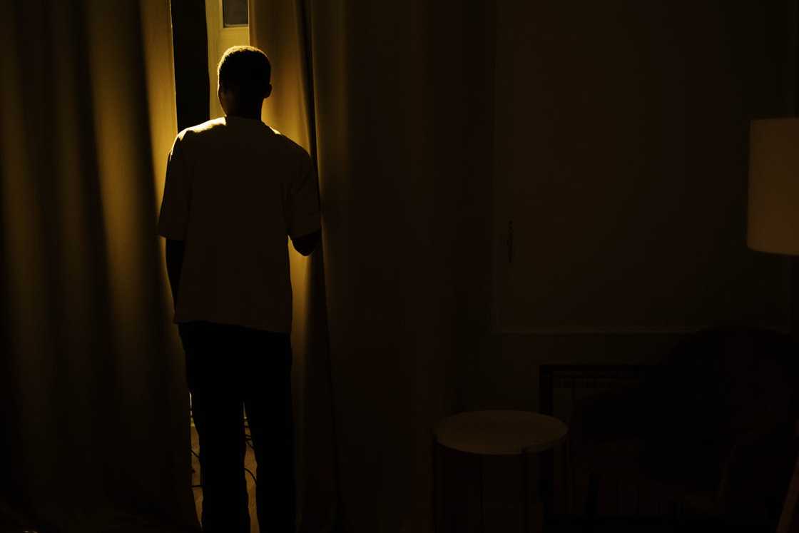 A man stands silently in a doorway between a dim living room and a lit kitchen.