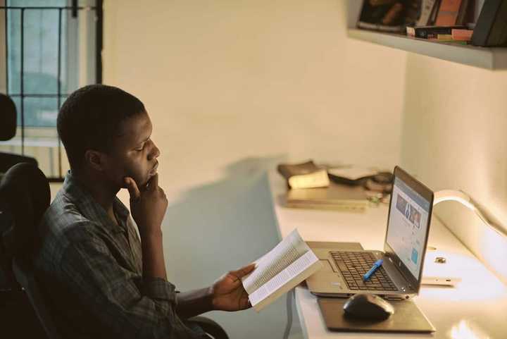 A young man in a checkered shirt is reading a book in front of a laptop